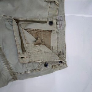 May include: Close-up of khaki-colored pants with a zipper and button closure. The pants have a faded appearance and visible stitching details. A label is visible on the back, and the text "GET YOUR K" is partially visible.