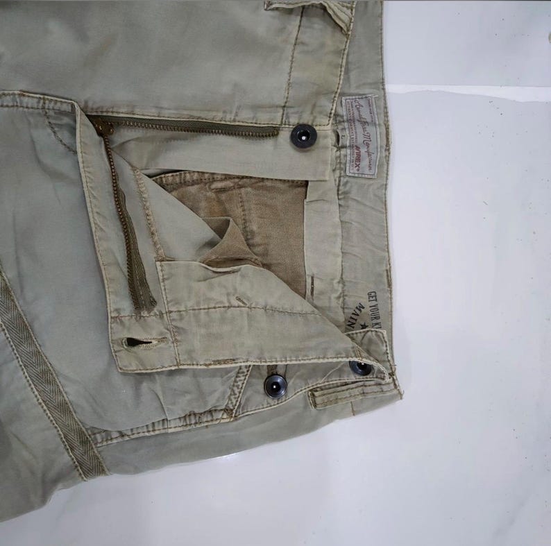 May include: Close-up of khaki-colored pants with a zipper and button closure. The pants have a faded appearance and visible stitching details. A label is visible on the back, and the text "GET YOUR K" is partially visible.