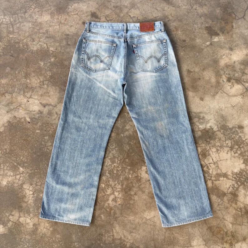 May include: Light blue denim jeans with a faded wash. The jeans have a classic five-pocket design, a red tag, and a leather patch. The jeans are laid flat on a concrete surface.