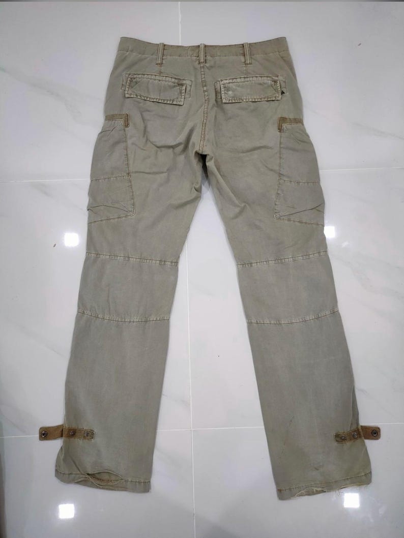 May include: Khaki-colored cargo pants with multiple pockets and reinforced stitching. Features include two back pockets, two side pockets, and decorative straps at the ankles. The pants are made of a durable fabric.