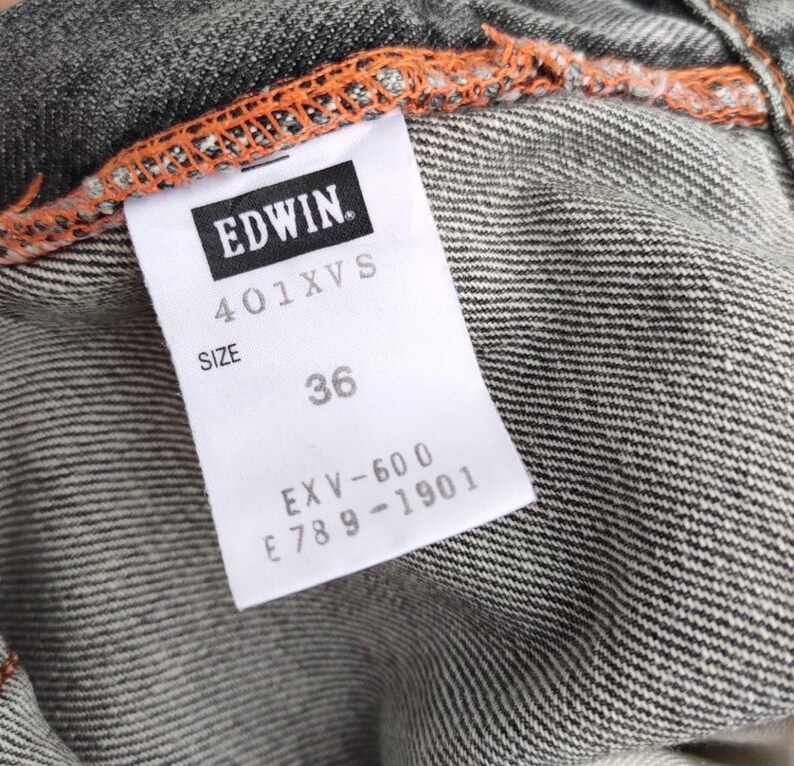 K&ouml;nnte beinhalten: Nahaufnahme eines Jeans-Kleidungsst&uuml;ck-Etiketts. Das wei&szlig;e Etikett zeigt die Marke "EDWIN" und die Gr&ouml;&szlig;e "36". Der Jeansstoff ist grau mit sichtbaren orangefarbenen N&auml;hten.