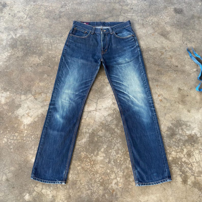 May include: Blue denim jeans with a classic five-pocket design. The jeans have a faded wash and a straight leg cut. The denim is a medium weight, and the jeans have a button and zipper closure. The jeans are laid flat on a concrete surface.