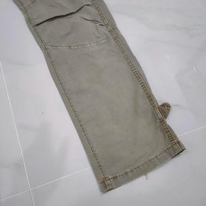 May include: Close-up of a section of olive green cargo pants. The pants have reinforced stitching, a knee patch, and a small loop detail. The fabric appears worn, with subtle variations in color.