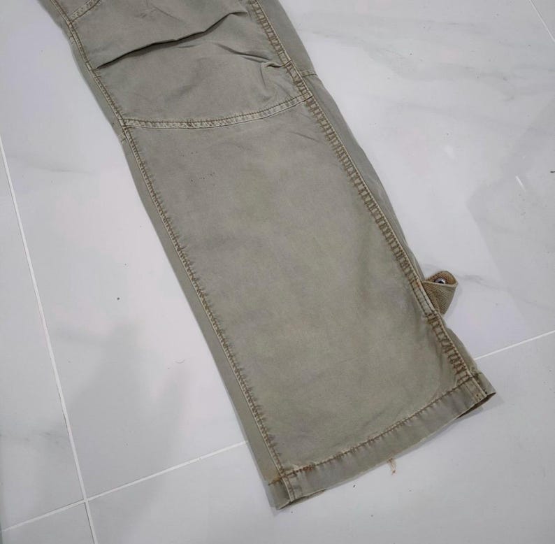 May include: Close-up of a section of olive green cargo pants. The pants have reinforced stitching, a knee patch, and a small loop detail. The fabric appears worn, with subtle variations in color.