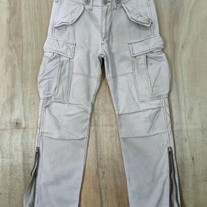 May include: Light gray cargo pants with multiple pockets and zippers on the lower legs. The pants are hanging on a hanger against a light wood background. The stitching is visible, and the pants appear to be made of a durable fabric.