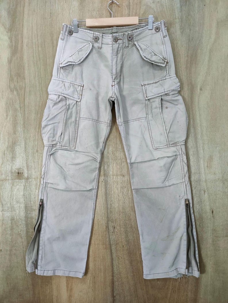 May include: Light gray cargo pants with multiple pockets and zippers on the lower legs. The pants are hanging on a hanger against a light wood background. The stitching is visible, and the pants appear to be made of a durable fabric.