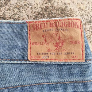 May include: Close-up of light blue denim fabric with a brown leather patch. The patch displays "TRUE RELIGION" and a guitar-playing figure. Other text includes "BRAND JEANS", "WORLD TOUR", "FASHION FOR THE SENSES", "SECTION JOEY", and "SEAT".