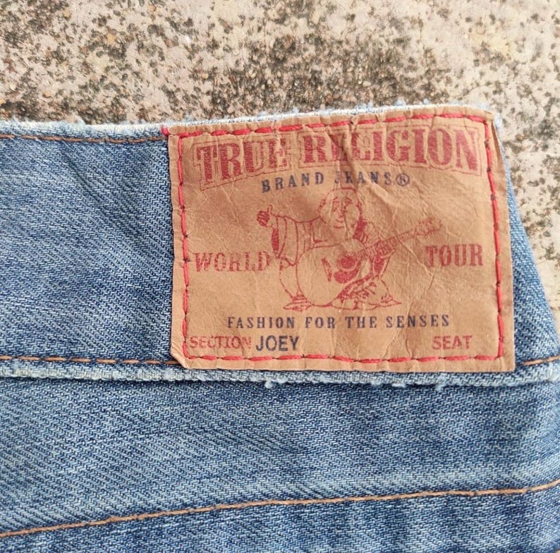 May include: Close-up of light blue denim fabric with a brown leather patch. The patch displays "TRUE RELIGION" and a guitar-playing figure. Other text includes "BRAND JEANS", "WORLD TOUR", "FASHION FOR THE SENSES", "SECTION JOEY", and "SEAT".