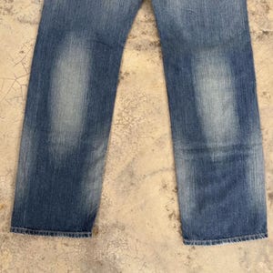 May include: Faded blue denim jeans with a straight leg cut. The jeans show wear and tear with lighter patches on the front of the legs. The bottom hems are frayed.