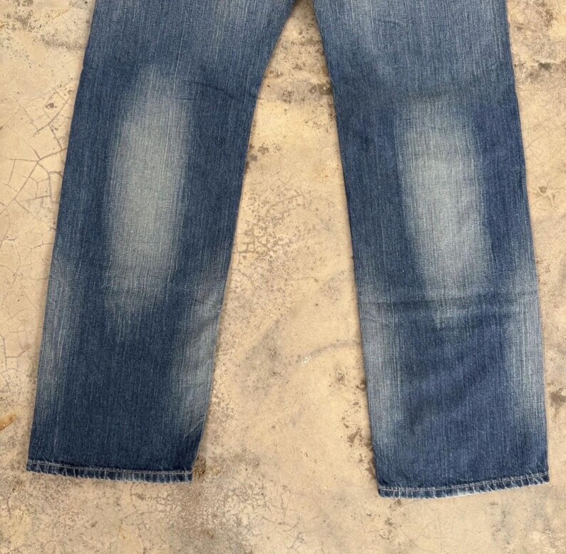 May include: Faded blue denim jeans with a straight leg cut. The jeans show wear and tear with lighter patches on the front of the legs. The bottom hems are frayed.