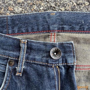 Pu&ograve; includere: Primo piano di jeans in denim blu scuro. I jeans presentano una chiusura con bottoni, una cerniera e dettagli di cucitura rossi. Il tessuto ha un aspetto usurato, con cuciture visibili e un bottone in metallo.