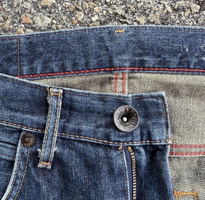 Pu&ograve; includere: Primo piano di jeans in denim blu scuro. I jeans presentano una chiusura con bottoni, una cerniera e dettagli di cucitura rossi. Il tessuto ha un aspetto usurato, con cuciture visibili e un bottone in metallo.