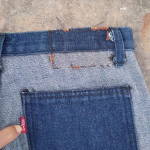 May include: Close-up of denim jeans with a two-tone design. The upper portion and back pocket are a darker blue, while the main body is a lighter blue. A red tag with white text is visible on the back pocket.