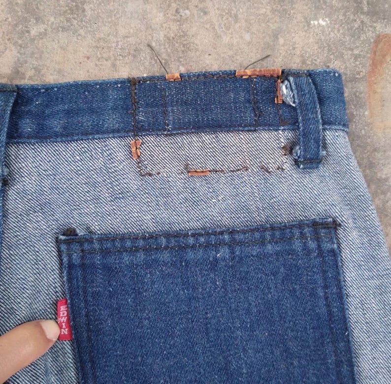 May include: Close-up of denim jeans with a two-tone design. The upper portion and back pocket are a darker blue, while the main body is a lighter blue. A red tag with white text is visible on the back pocket.