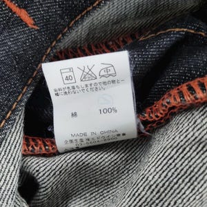 Pu&ograve; includere: Primo piano di tessuto denim scuro con cuciture arancioni. Un'etichetta bianca mostra le istruzioni di lavaggio, tra cui un simbolo di lavaggio a 40 gradi, e il testo "100%" e "MADE IN CHINA".