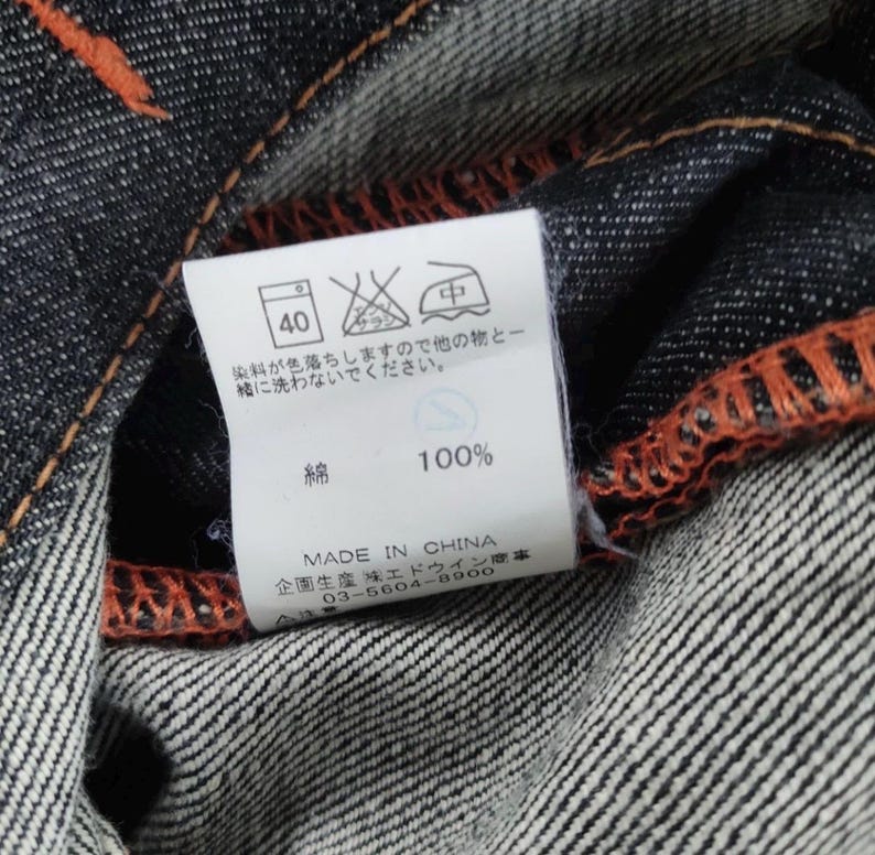 Pu&ograve; includere: Primo piano di tessuto denim scuro con cuciture arancioni. Un'etichetta bianca mostra le istruzioni di lavaggio, tra cui un simbolo di lavaggio a 40 gradi, e il testo "100%" e "MADE IN CHINA".