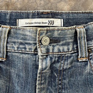 Puede incluir: Primer plano de unos vaqueros de mezclilla azules con cierre de bot&oacute;n y una etiqueta que dice "Exclusive Vintage Model XU". Los vaqueros tienen un lavado descolorido y costuras visibles.