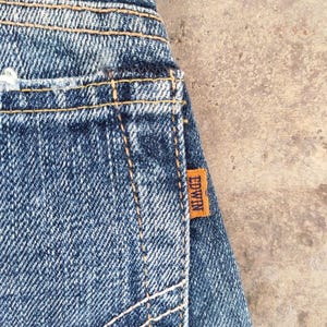May include: Close-up of blue denim jeans with visible stitching and an orange Edwin brand tag. The jeans have a worn appearance, with faded areas and a distressed look.