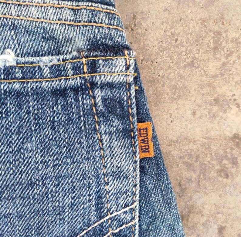 May include: Close-up of blue denim jeans with visible stitching and an orange Edwin brand tag. The jeans have a worn appearance, with faded areas and a distressed look.