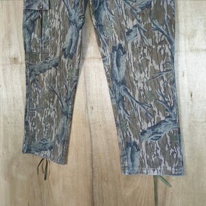 May include: Camouflage cargo trousers in shades of green, brown, and blue. The trousers feature a tree bark pattern, two cargo pockets, and drawstrings at the ankles. The fabric appears to be a durable cotton blend.