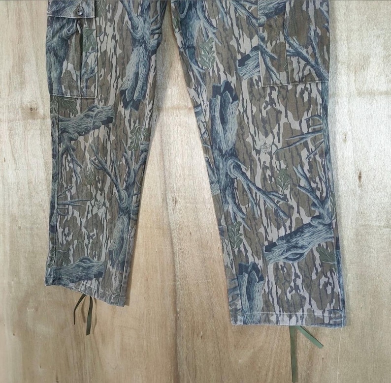 May include: Camouflage cargo trousers in shades of green, brown, and blue. The trousers feature a tree bark pattern, two cargo pockets, and drawstrings at the ankles. The fabric appears to be a durable cotton blend.