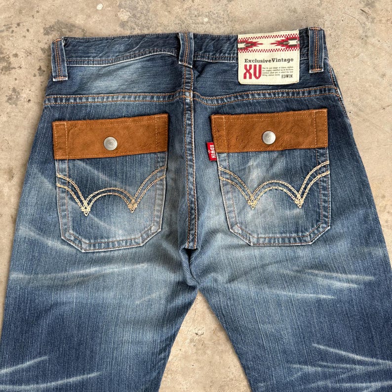 Op de afbeelding: Blauwe spijkerbroek met bruine su&egrave;de zakaccenten en een knoopsluiting. Voorzien van het "Exclusive Vintage" label en "XU" branding. De achterzakken hebben een uniek stikselontwerp.