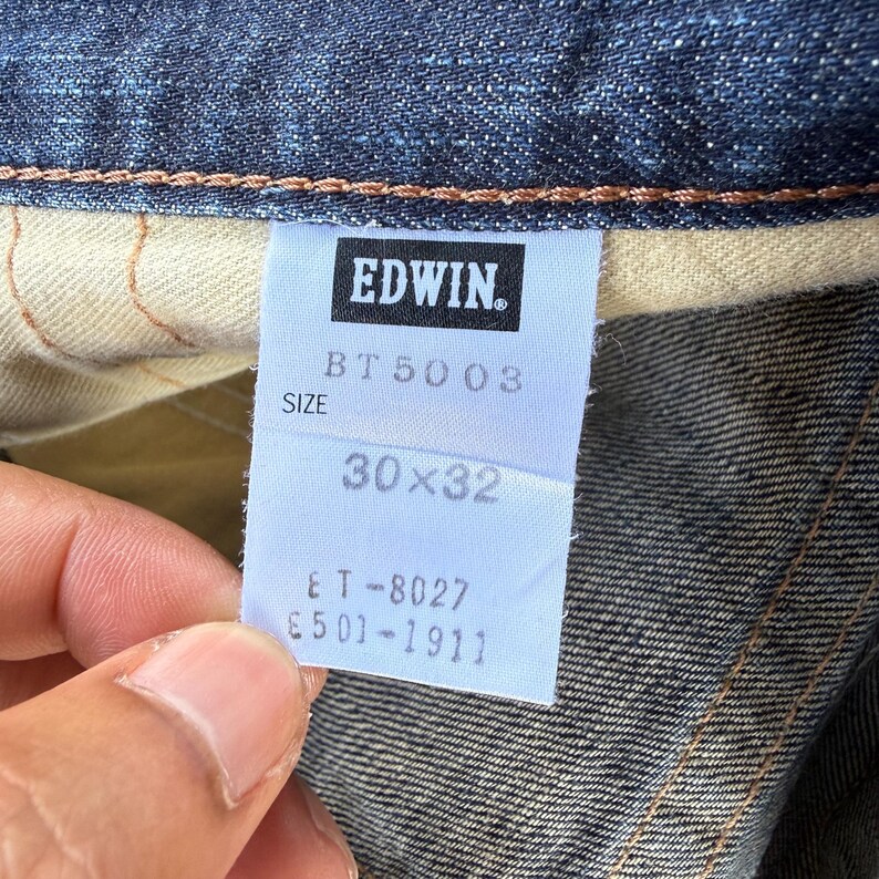 May include: Close-up of a denim jeans label with the brand name "EDWIN" in a black rectangle. The label indicates size 30x32. The jeans are a dark blue color.