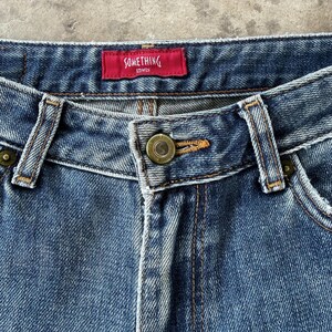 May include: Close-up of blue denim jeans with a button closure and a red tag that reads "SOMETHING EDWIN". The jeans have a classic five-pocket design and are made of durable denim fabric.