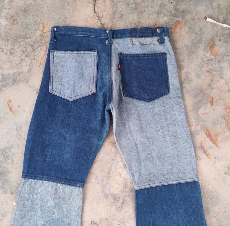 May include: Two-tone denim jeans with a patchwork design. The jeans feature a mix of light and dark blue denim panels, with a classic five-pocket design. The back pockets are visible, and a red tag is on one pocket.