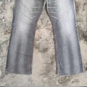 K&ouml;nnte beinhalten: Verblasste graue Jeans mit ausgestelltem Bein. Die Jeans haben ein klassisches F&uuml;nf-Taschen-Design mit sichtbaren N&auml;hten in einem kontrastierenden braunen Faden entlang der N&auml;hte und Taschen. Der Denim hat ein abgenutztes, leicht verwaschenes Aussehen.