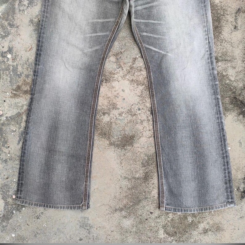 K&ouml;nnte beinhalten: Verblasste graue Jeans mit ausgestelltem Bein. Die Jeans haben ein klassisches F&uuml;nf-Taschen-Design mit sichtbaren N&auml;hten in einem kontrastierenden braunen Faden entlang der N&auml;hte und Taschen. Der Denim hat ein abgenutztes, leicht verwaschenes Aussehen.