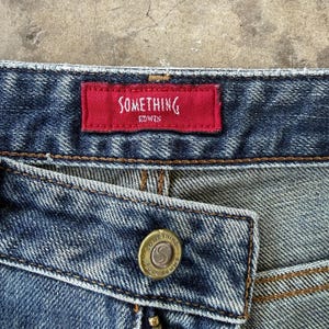 May include: Close-up of denim jeans with a red tag that reads "SOMETHING EDWIN" in white lettering. The jeans have a button with the words "SOMETHING POUR FEMME" around a central design.