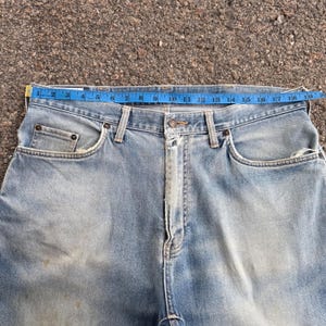 May include: Light blue denim jeans with a blue measuring tape across the waist. The jeans have a classic five-pocket design, with visible wear and tear. The waist measures approximately 18 inches across.