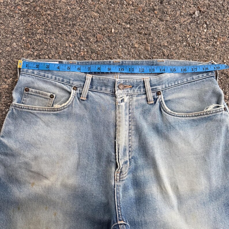 May include: Light blue denim jeans with a blue measuring tape across the waist. The jeans have a classic five-pocket design, with visible wear and tear. The waist measures approximately 18 inches across.