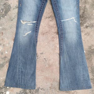 May include: Light blue denim jeans with a flared leg design. The jeans feature distressed details, including rips on the knees. The fabric shows signs of wear and fading, giving them a vintage look. A classic style of jeans.