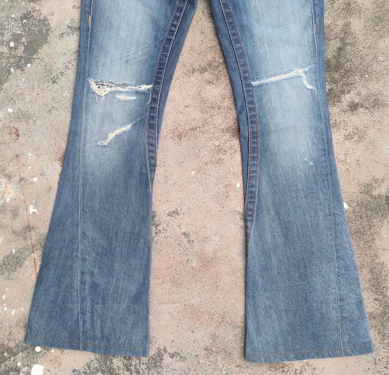 May include: Light blue denim jeans with a flared leg design. The jeans feature distressed details, including rips on the knees. The fabric shows signs of wear and fading, giving them a vintage look. A classic style of jeans.