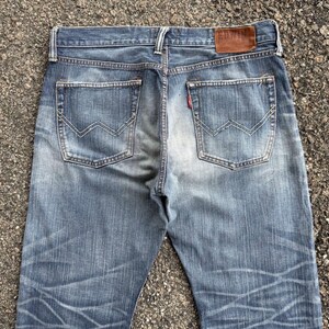 May include: Faded blue denim jeans with a classic five-pocket design. Features include a brown leather patch with the brand name "EDWIN" and a red tab on the back pocket. The jeans have a worn, distressed look.