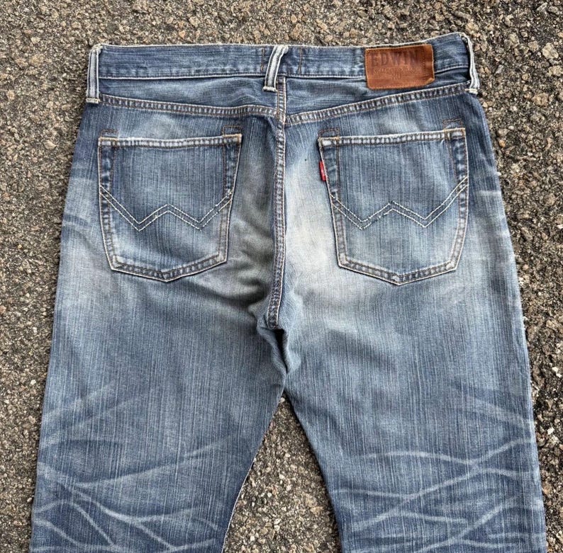 May include: Faded blue denim jeans with a classic five-pocket design. Features include a brown leather patch with the brand name "EDWIN" and a red tab on the back pocket. The jeans have a worn, distressed look.