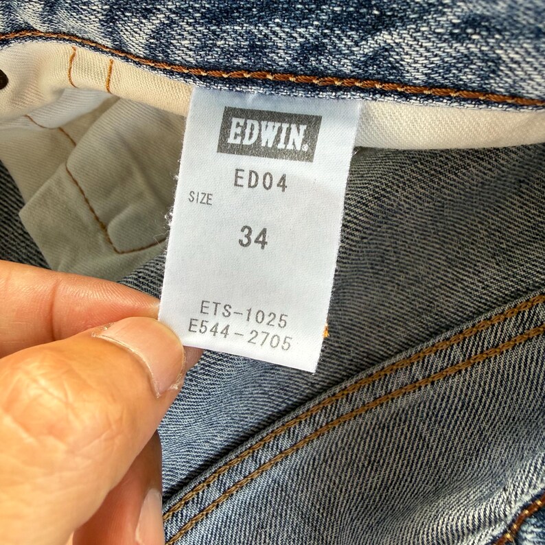 Pu&ograve; includere: Primo piano di un'etichetta di jeans in denim con il marchio "EDWIN" in un riquadro rettangolare. L'etichetta mostra la taglia "34" e il codice stile "ED04". L'etichetta include anche i codici "ETS-1025" e "E544-2705".