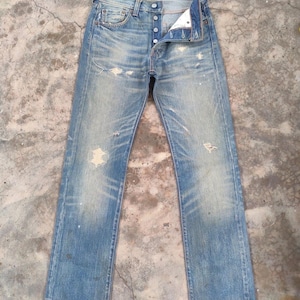 Puede incluir: Vaqueros azules descoloridos con bragueta de botones y detalles desgastados. Los vaqueros tienen un aspecto desgastado con desgaste visible, incluyendo rasgaduras y decoloración. El diseño clásico de cinco bolsillos es visible.