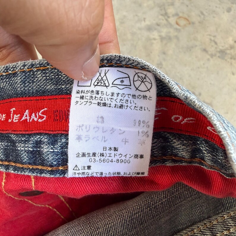 May include: Close-up of a denim jeans label with Japanese text and care instructions. The label is white with black text, and the jeans have a red interior lining. The label includes fabric composition details.