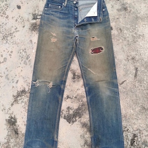 Könnte beinhalten: Verblasste blaue Jeans mit abgenutzten Details, einschließlich Rissen und Flicken. Die Jeans haben einen Knopfverschluss und ein klassisches Fünf-Taschen-Design. Der Markenname "A.P.C." ist auf der Gesäßtasche sichtbar.