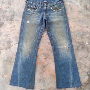 May include: Faded blue denim jeans with a flared leg design. The jeans show signs of wear, including distressed areas and a small hole on the right leg. Red stitching accents the seams.