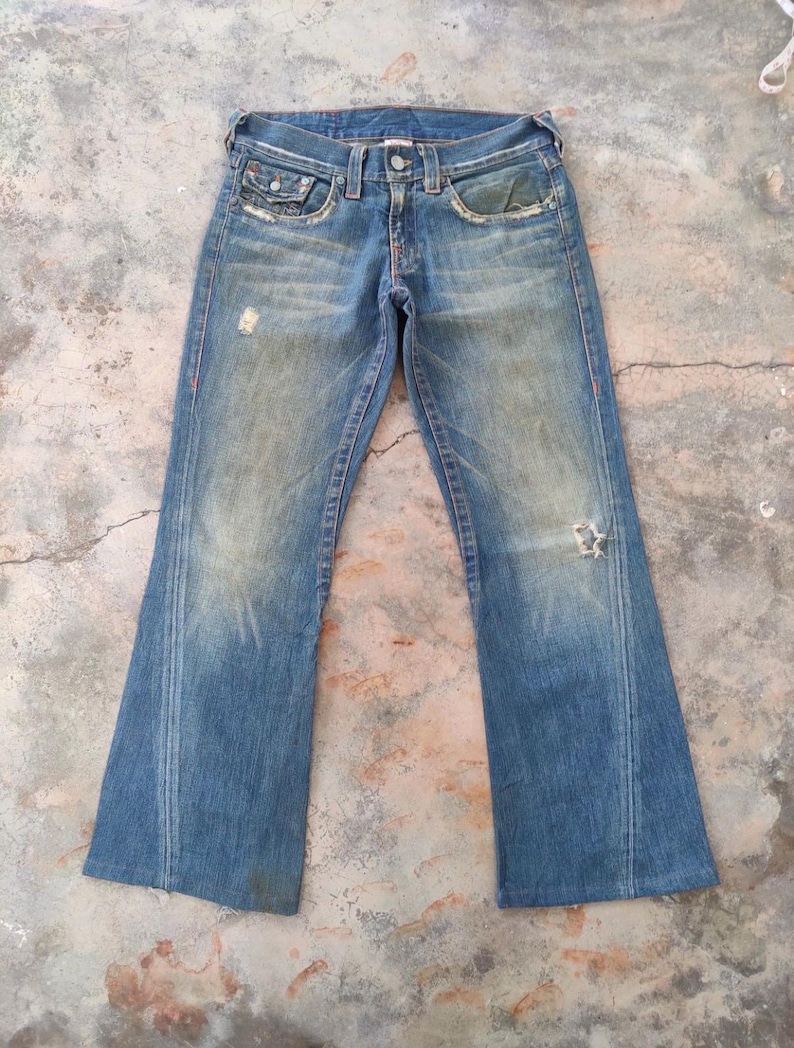 May include: Faded blue denim jeans with a flared leg design. The jeans show signs of wear, including distressed areas and a small hole on the right leg. Red stitching accents the seams.