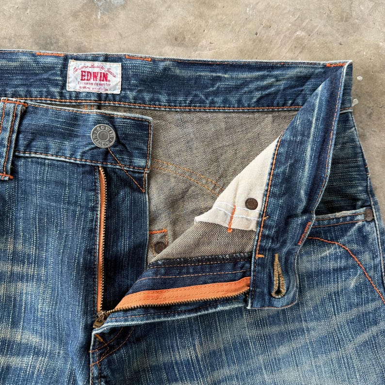 Pu&ograve; includere: Primo piano di jeans di denim blu con patta aperta. I jeans hanno una cerniera e cuciture arancioni, con chiusura a bottone. Il marchio "EDWIN" &egrave; visibile su un'etichetta. Il denim ha un aspetto usurato e sbiadito.