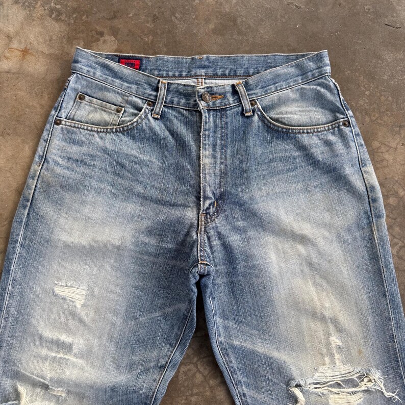 May include: Light blue denim jeans with distressed details, including rips and faded areas. The jeans have a classic five-pocket design, a button and zipper closure, and a red tag on the back.