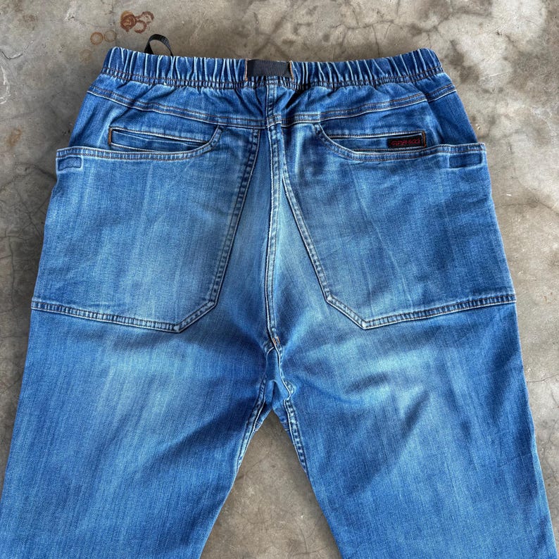 May include: Back view of blue denim pants with an elastic waistband and two back pockets. The pants have a faded wash and a small logo patch on the back pocket. The stitching is visible throughout the design.