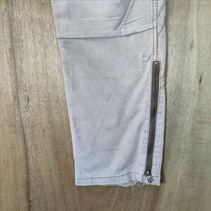 May include: Close-up of light gray pants with a side zipper and distressed hem. The pants have visible staining and are made of a durable fabric. The zipper is a dark brown color.