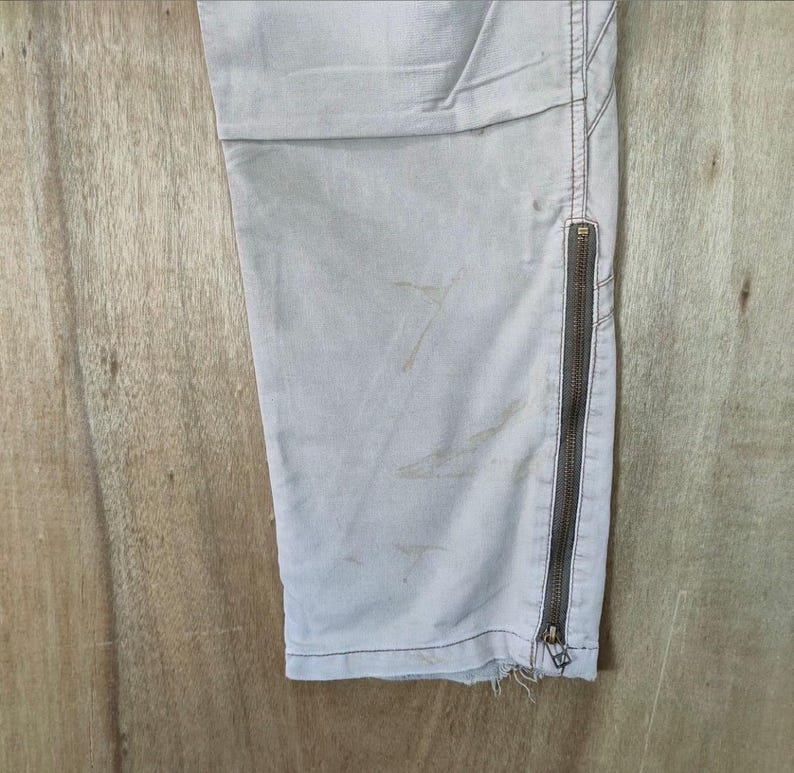 May include: Close-up of light gray pants with a side zipper and distressed hem. The pants have visible staining and are made of a durable fabric. The zipper is a dark brown color.