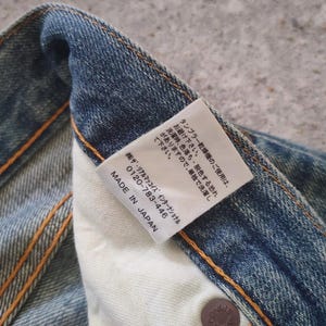 May include: Close-up of a section of blue denim jeans with visible stitching and a white tag. The tag has Japanese text and the words "MADE IN JAPAN". The jeans have a worn appearance.